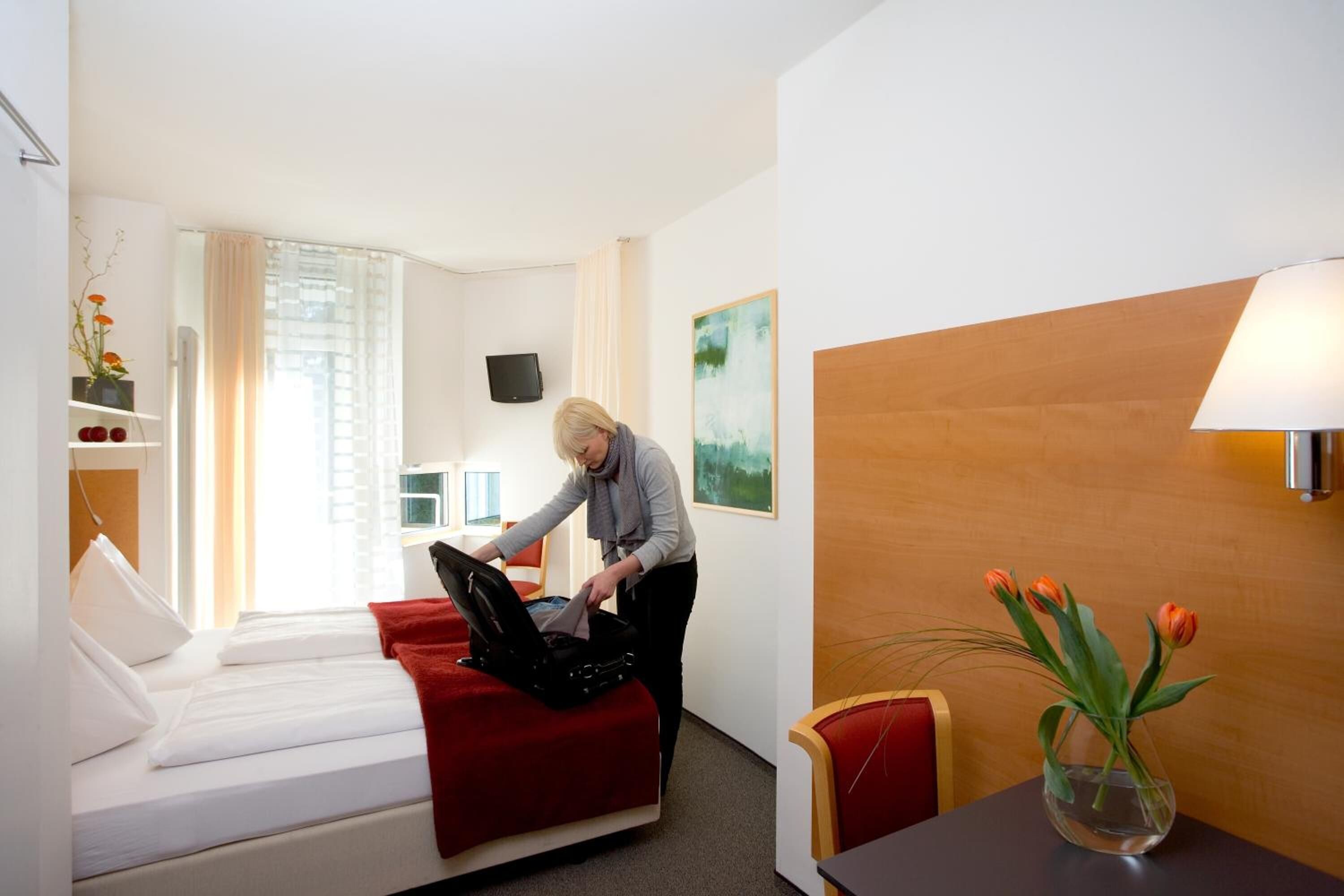 Una habitación de hotel con una cama, un sofá rojo, un escritorio con una lámpara, un jarrón con flores y un cuadro en la pared.