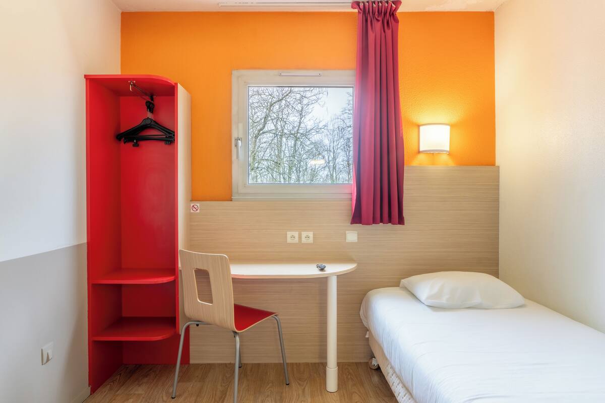 Een kleine hotelkamer met een enkel bed, een bureau, een stoel, een kledingkast en een raam met uitzicht op bomen.