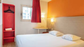 A hotel room with a bed, a red wardrobe, a small table, and a window with a curtain.