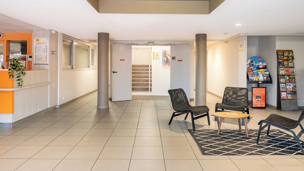 Eine Lobby mit einer Rezeption, zwei Sessel, einem kleinen Tisch, einem Zeitschriftenständer und einer Treppe.
