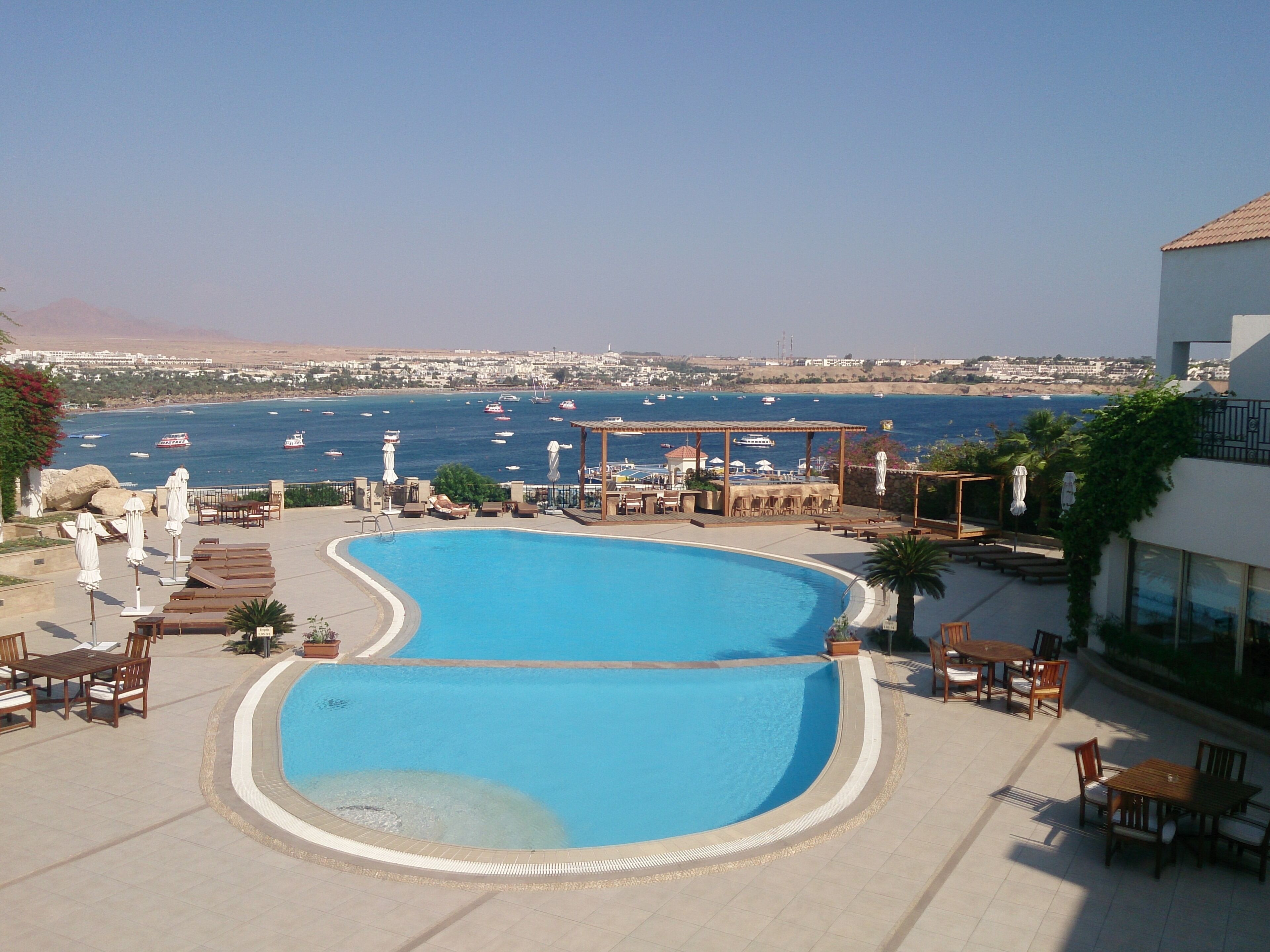 Una piscina con vistas al mar y a barcos, rodeada de sillones de baño y sombrillas.