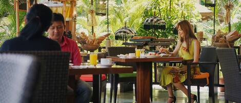 Un ristorante con posti a sedere all'aperto, tavoli in legno, sedie in vimini e un giardino tropicale sullo sfondo.