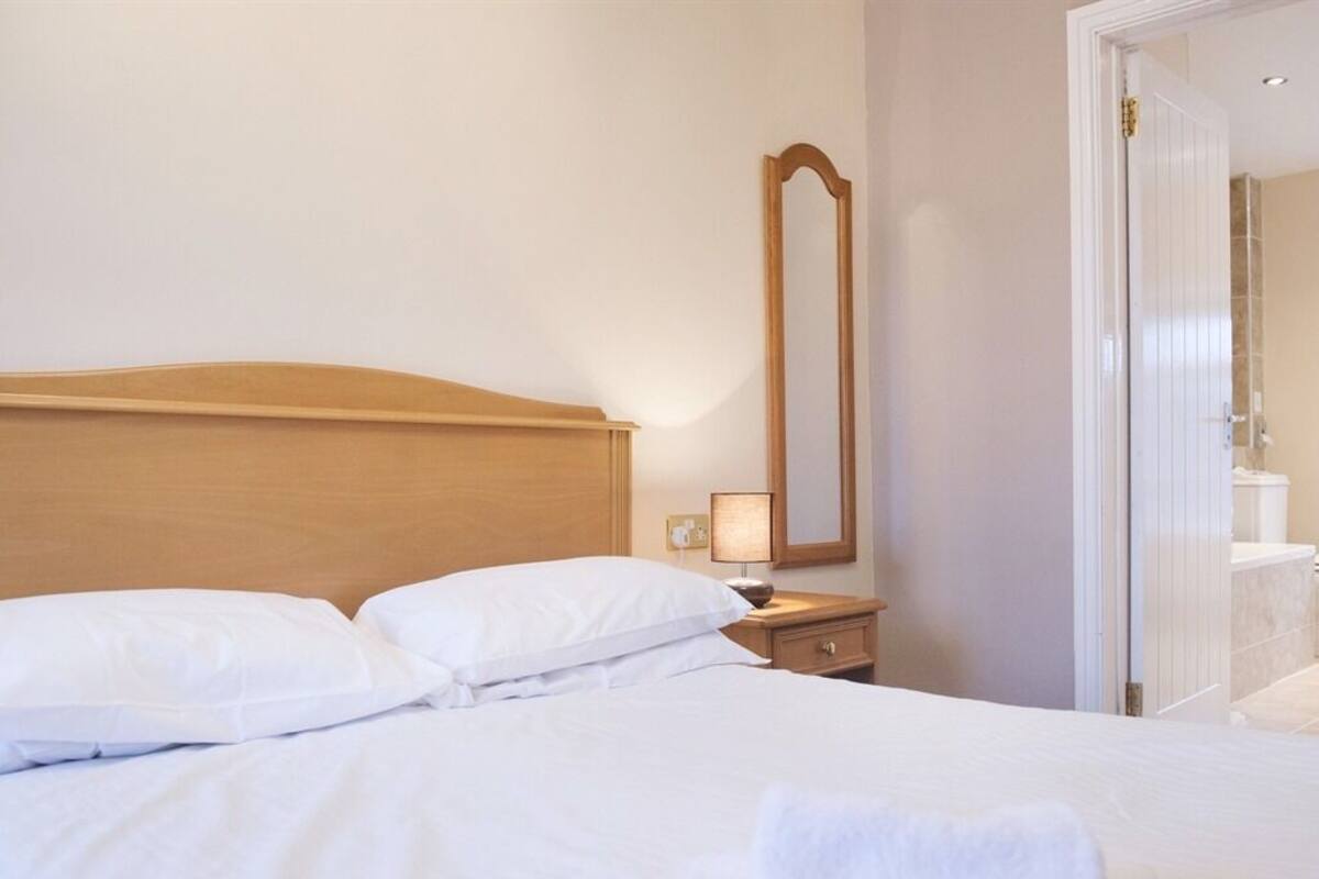 A neatly made bed with white linens and pillows, a wooden headboard, a bedside table with a lamp, and a full-length mirror on the wall.
