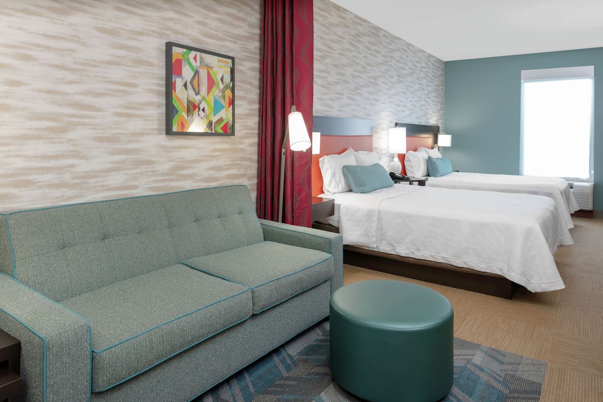 Ein Hotelzimmer mit einer Couch, zwei Betten, einem runden Hocker, einem großen Fenster und einem farbenfrohen, abstrakten Gemälde an der Wand.