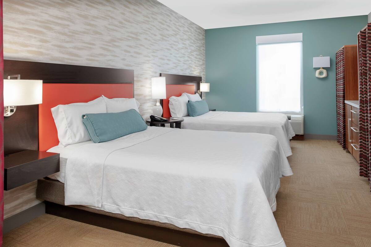 Ein Hotelzimmer mit zwei Betten, jeweils mit weißer Bettwäsche und türkisfarbenen Kissen, einer roten Akzentwand und einem Fenster mit Vorhängen.