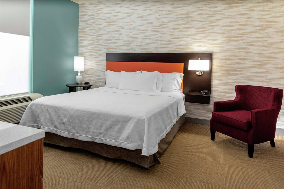 Ein Hotelzimmer mit einem Bett, einem Nachttisch mit Lampe, einem roten Sessel und einer strukturierten Wand.