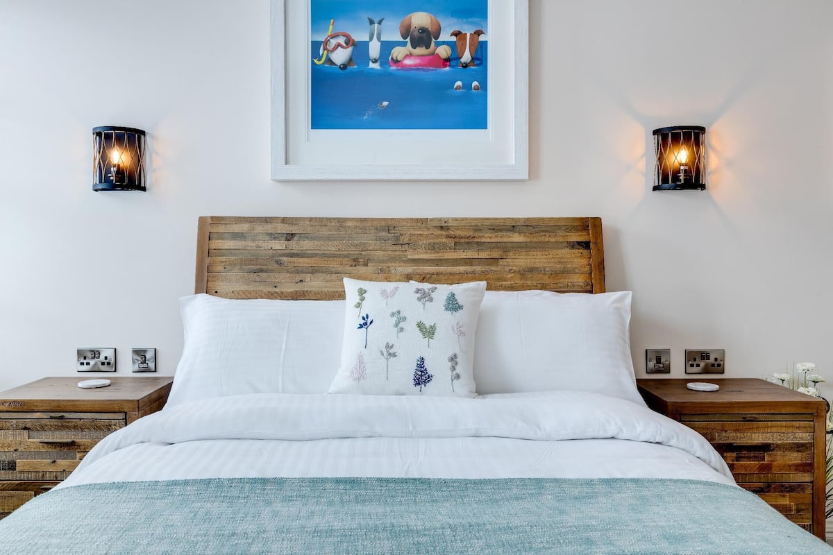 A neatly made bed with white linens and a blue throw blanket, flanked by wooden nightstands with wall sockets and framed artwork above.