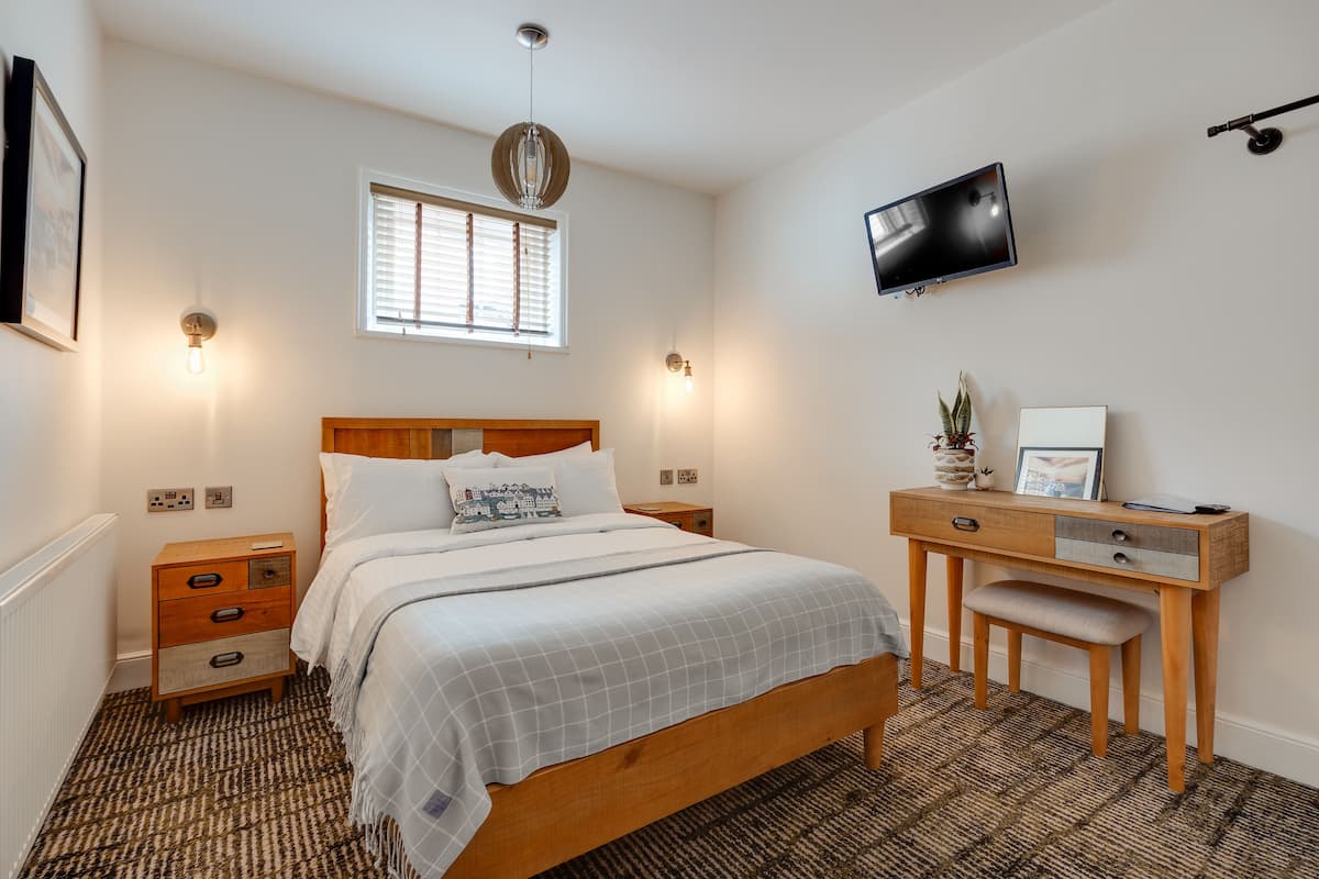 A bedroom with a bed, bedside tables, a desk with a chair, a TV, and a window with blinds.