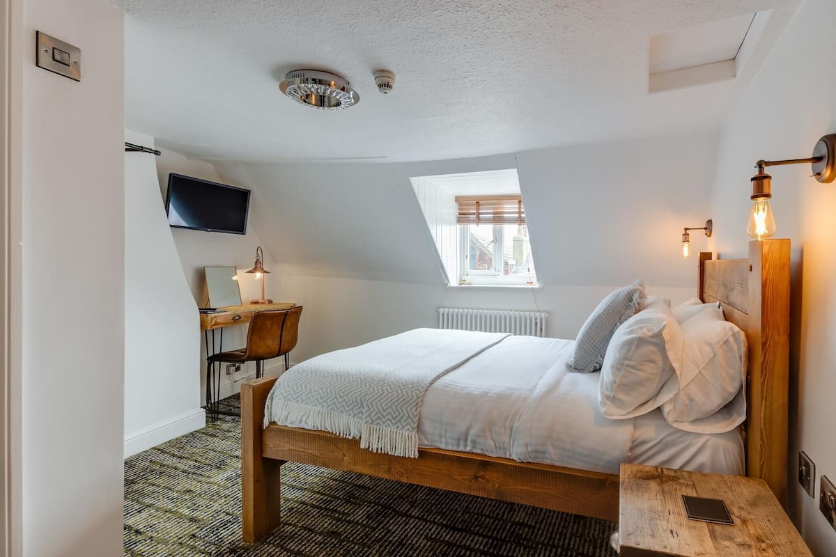 A bedroom with a wooden bed, a desk with a chair, a TV mounted on the wall, and a window with blinds.