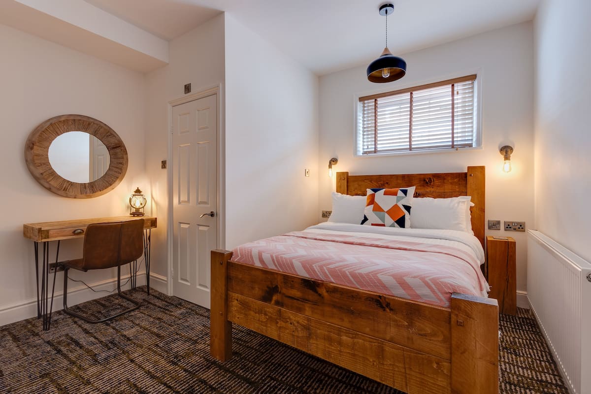 A bedroom with a wooden bed, a desk, a chair, a round mirror, and a window with blinds.