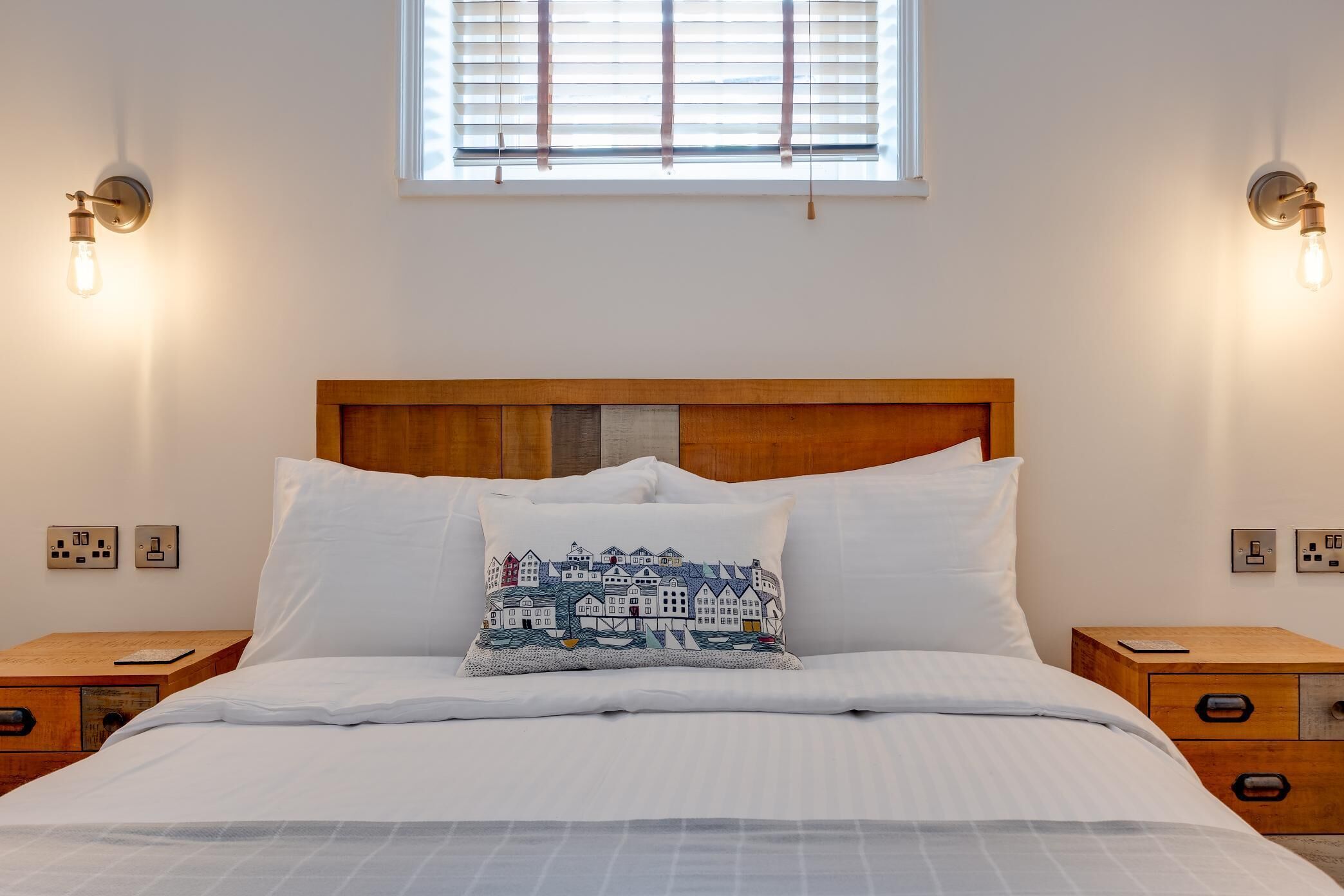 A neatly made bed with a white comforter and pillows, a wooden headboard, bedside tables with drawers, and wall-mounted light fixtures.
