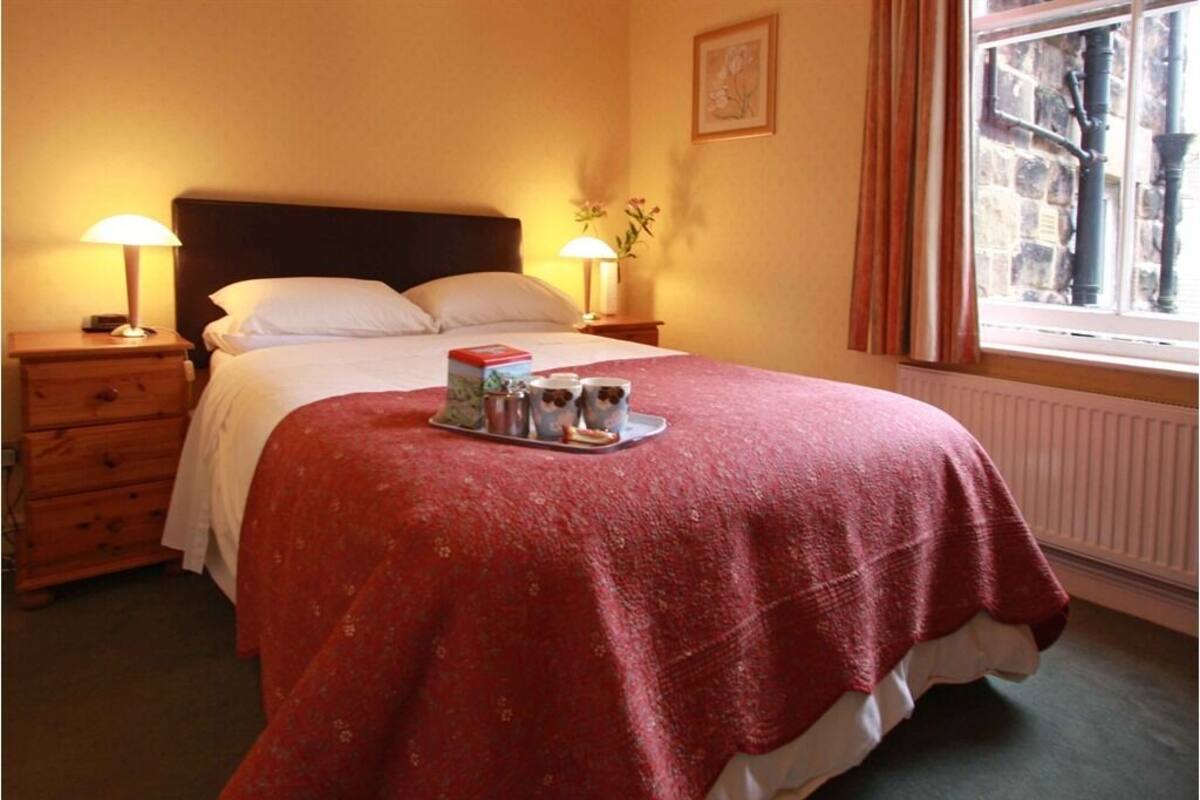 A neatly made bed with a red blanket, a tray with two mugs and a book, and a window with a view of the outside.