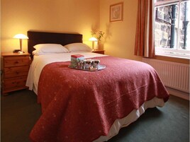 A neatly made bed with a red blanket, a tray with two mugs and a book, and a window with a view of the outside.