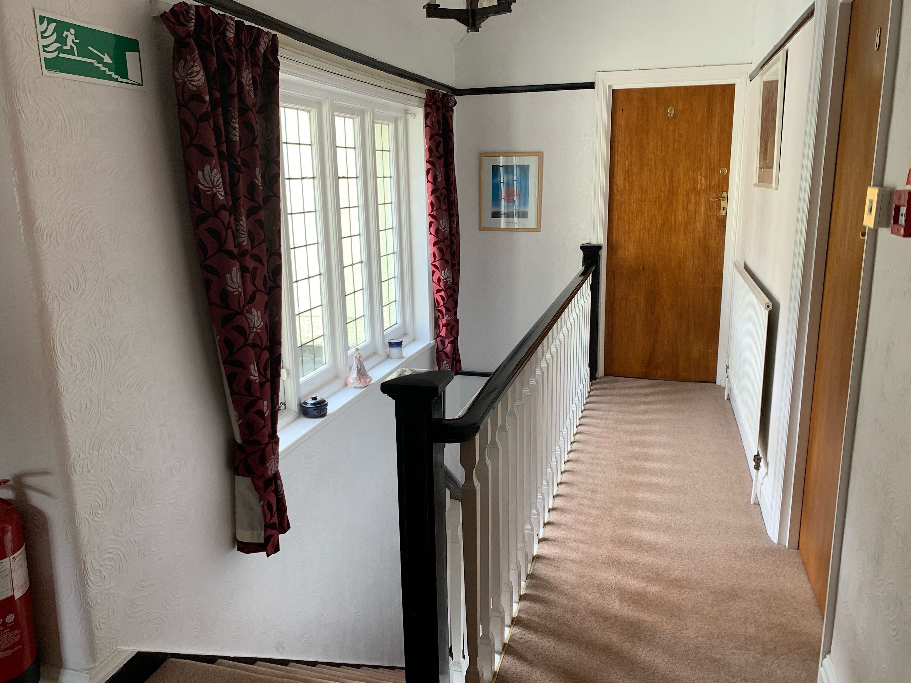 A hallway with a white wall, a window with red curtains, a framed picture, and a wooden door at the end.