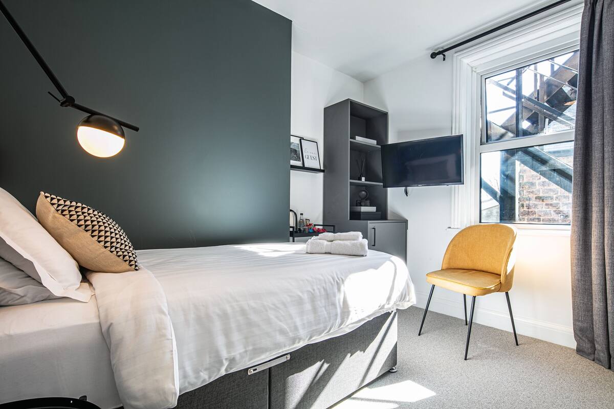 Ein modernes Schlafzimmer mit Bett, Sessel, Fernseher und Fenster mit Blick auf ein Gebäude.