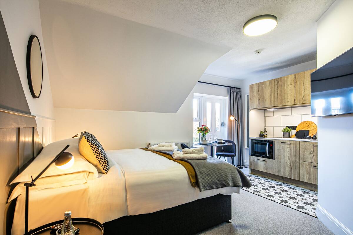 Ein modernes Hotelzimmer mit Bett, Nachttischlampe und einem kleinen Tisch mit einer Flasche und einer Fernbedienung. Im Hintergrund ist eine Küchenzeile mit Holzausstattung und einer Mikrowelle erkennbar.