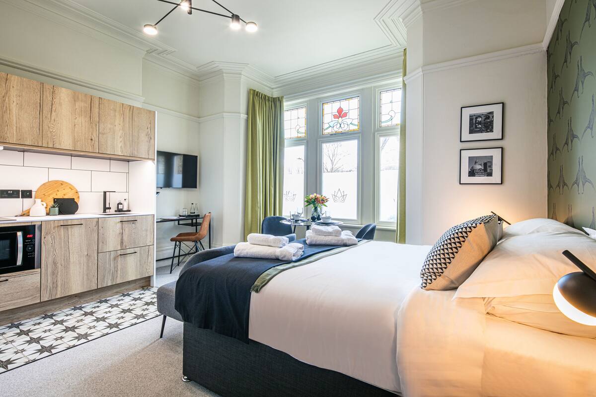 Ein modernes Hotelzimmer mit einem großen Bett, einer separaten Sitzecke, einer Küchenzeile und Blick ins Freie durch ein Fenster.