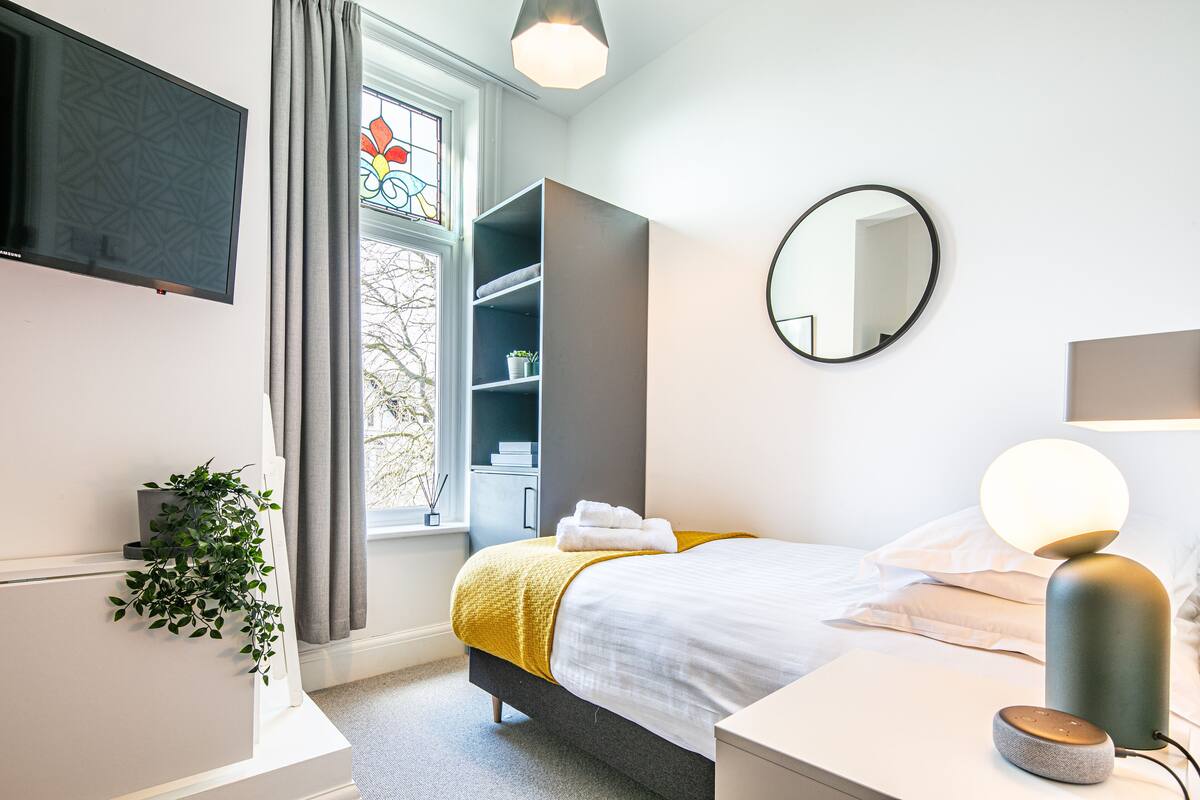 Ein modernes Schlafzimmer mit Bett, Nachttisch, Wand-Fernseher, rundem Spiegel und einem Fenster mit Vorhängen.