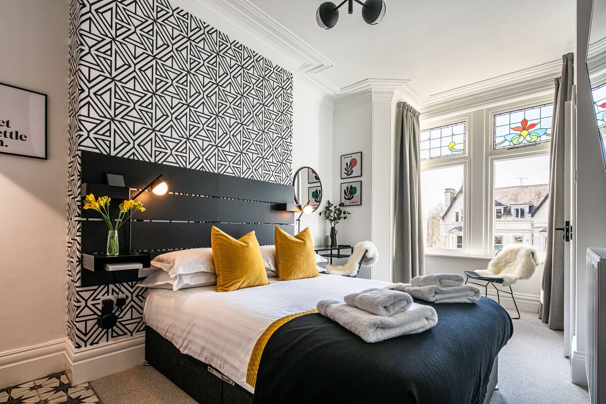 Ein modernes Hotelzimmer mit Wand in geometrischem Muster, einem Bett mit weißer und gelber Bettwäsche, einem Nachttisch mit Lampe und Blick auf die Stadt durch große Fenster.