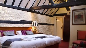 A bedroom with a large bed, a bedside table with a lamp, a red chair, and a door leading to another room. - The Saracens Head (Amersham)