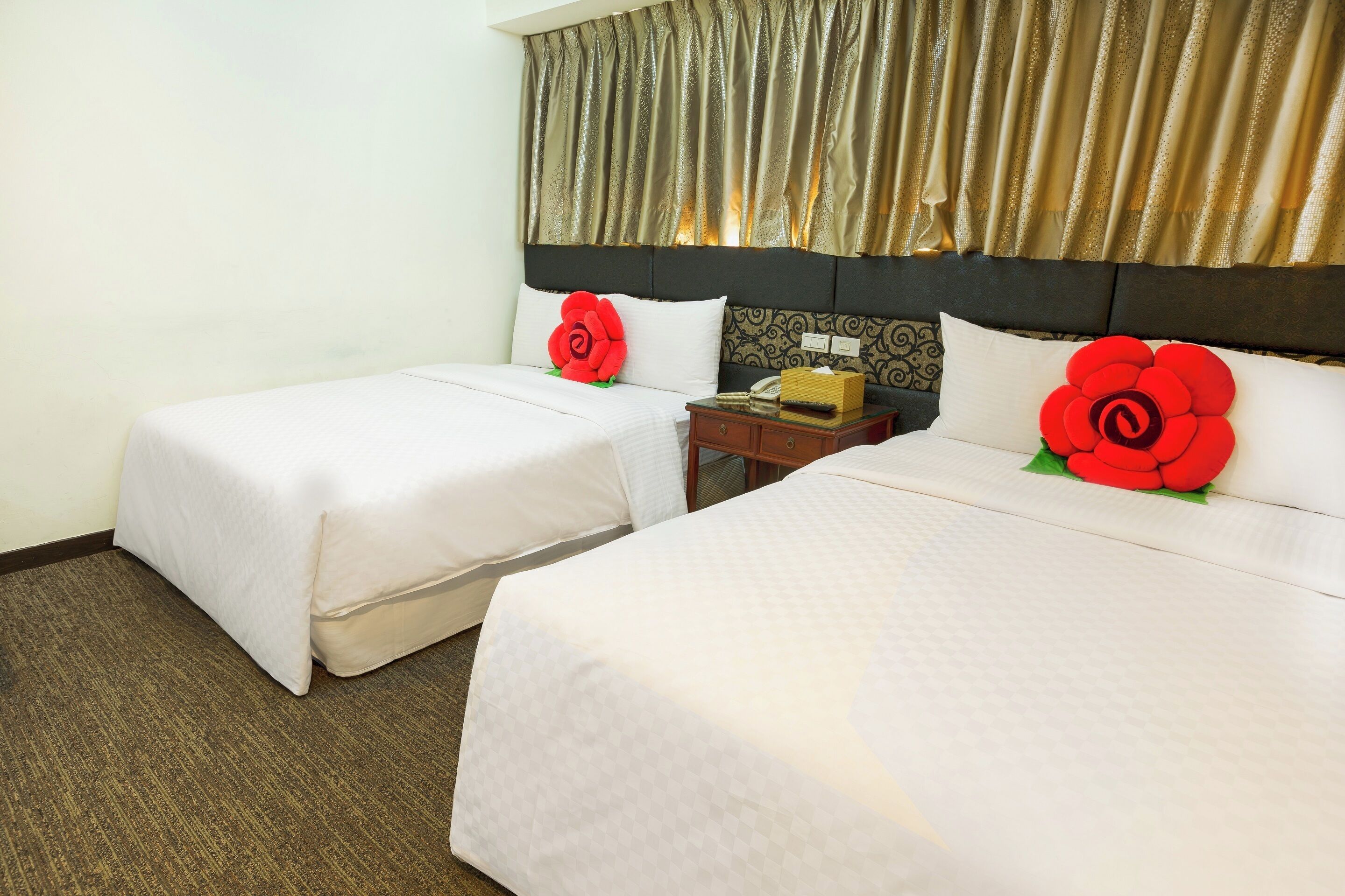 A hotel room with two beds, each with a white bedspread and a red floral pillow. There is a wooden nightstand with a telephone and a box between the beds. The room has a carpeted floor and a window with curtains.