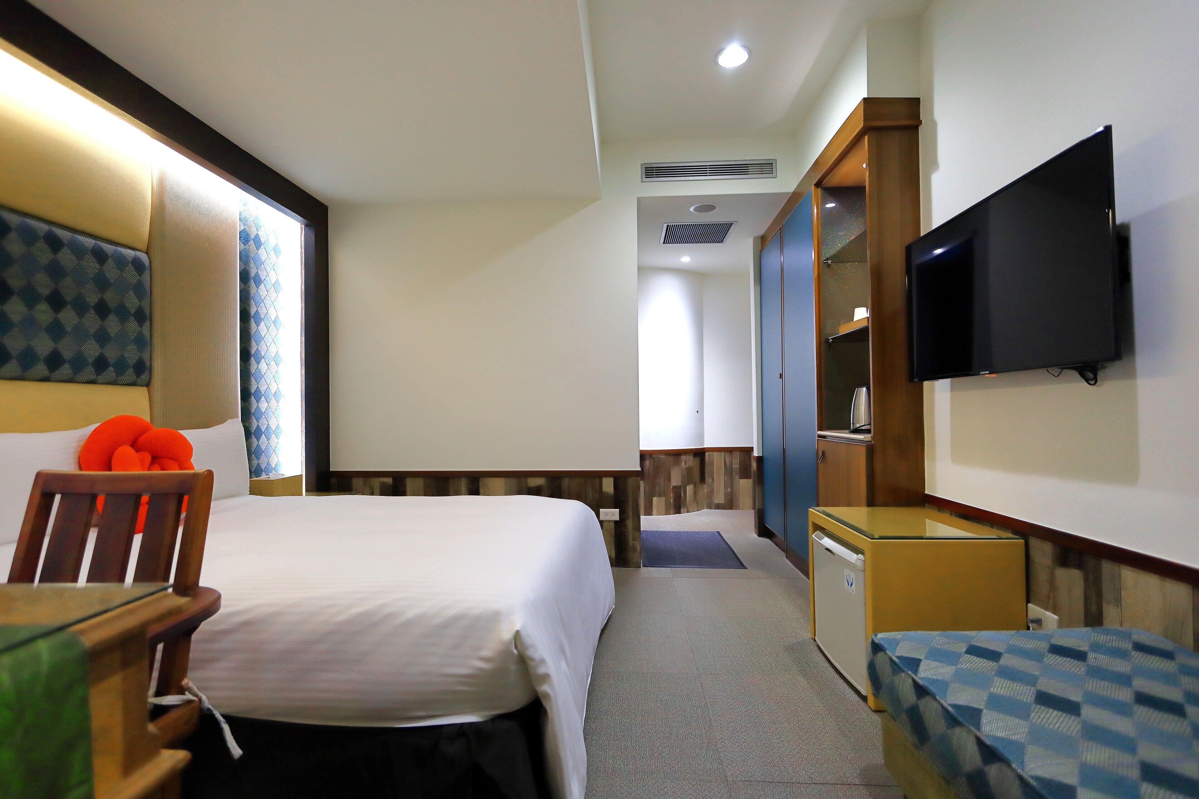 A hotel room with a bed, a chair, a small table, a TV mounted on the wall, and a mini-fridge.