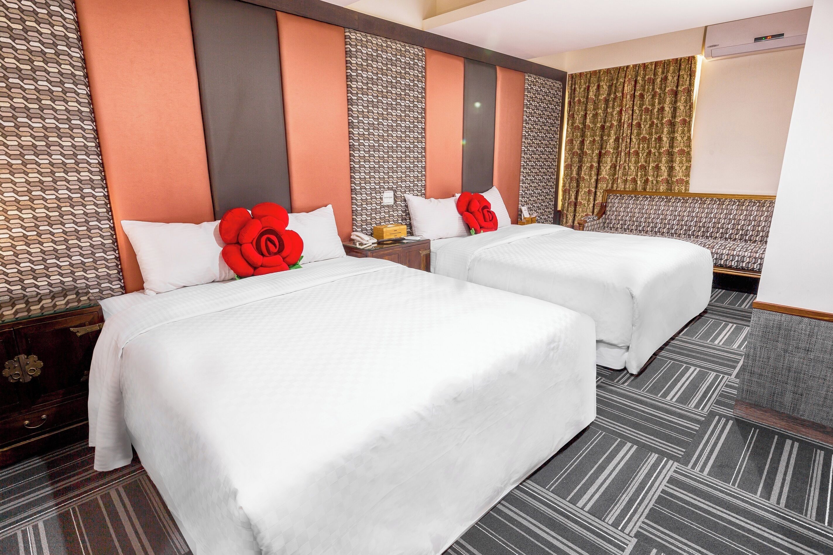 A hotel room with two beds, each with a white bedspread and red rose decorations. The room has a patterned wall and a large window with curtains.