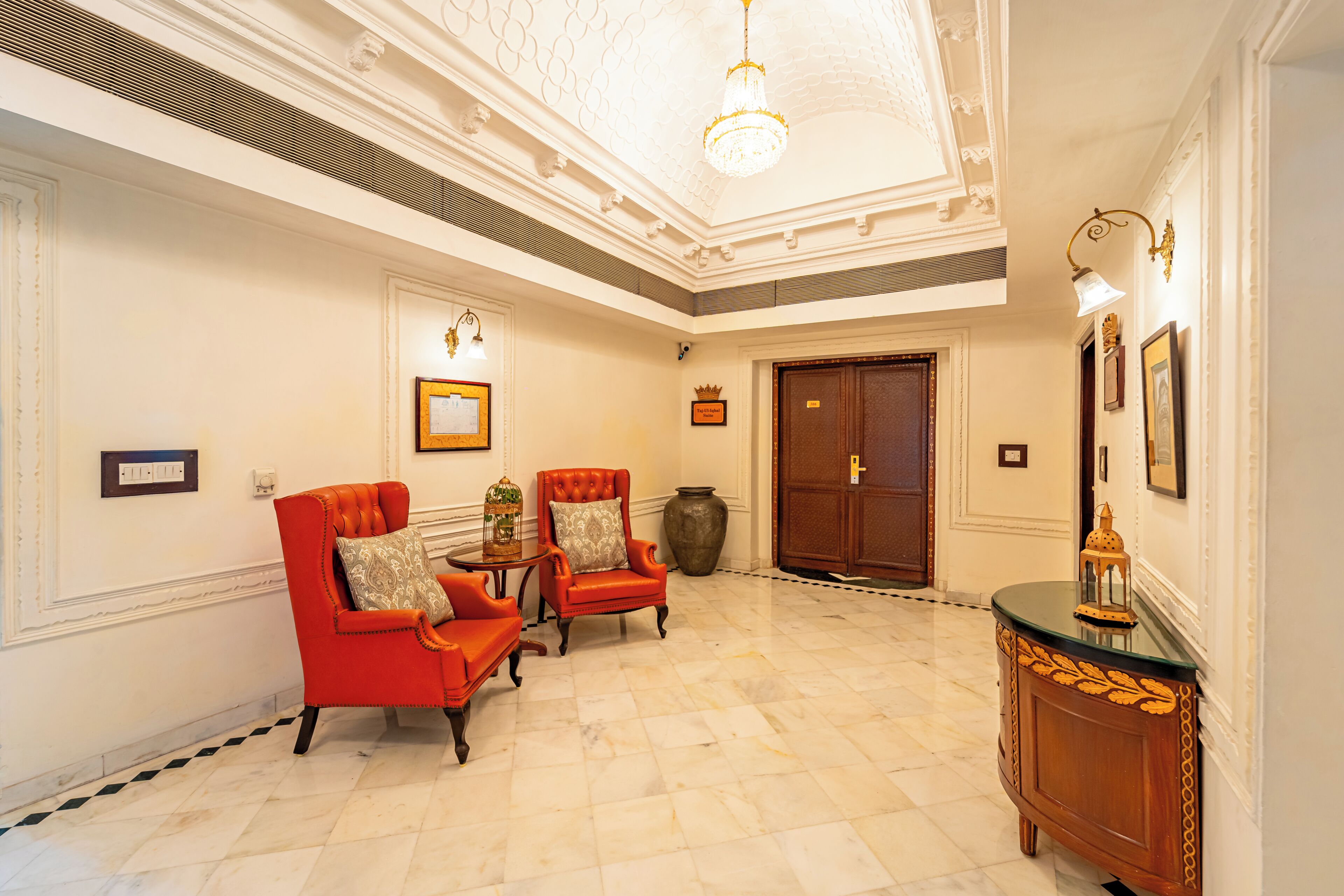 A spacious lobby with a high ceiling, a chandelier, two red armchairs, a wooden cabinet, and a large vase.