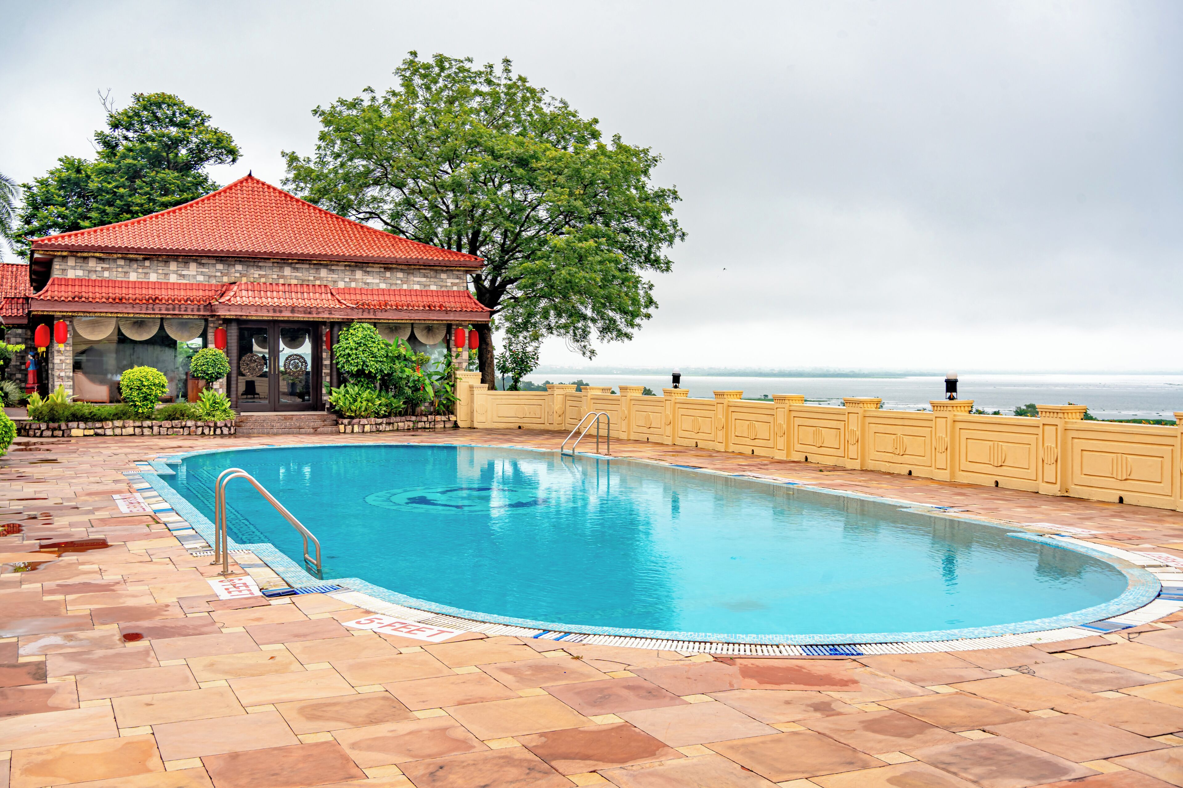 A swimming pool with a tiled deck, a building with a red roof, and a view of the sea.