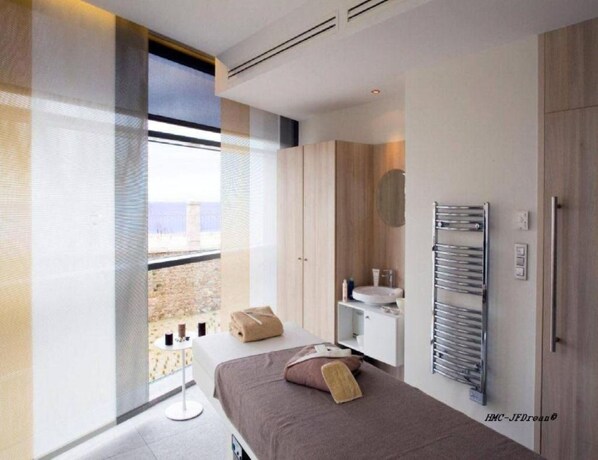 Una habitación de hotel moderna con un ventanal grande, una cama con manta marrón, una mesita de noche y un lavamanos integrado.