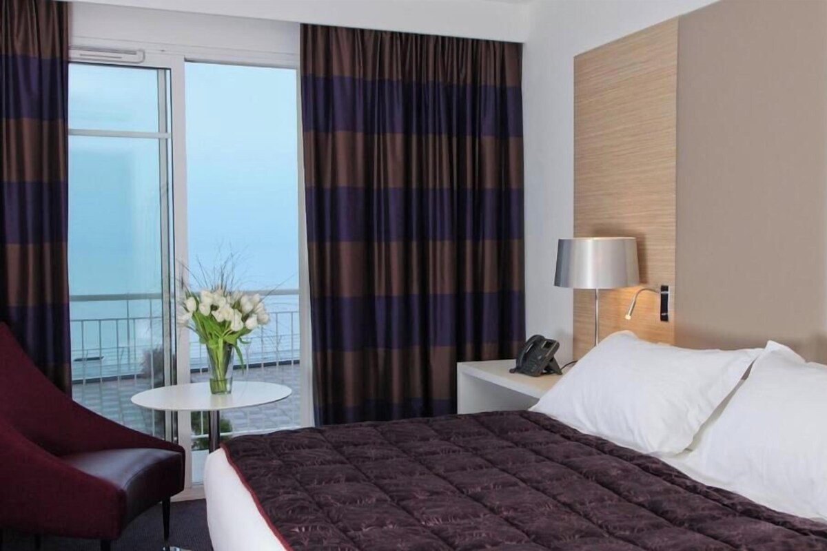A hotel room with a bed, a bedside table with a lamp, a chair, a small table with a vase of flowers, and a view of the outside.
