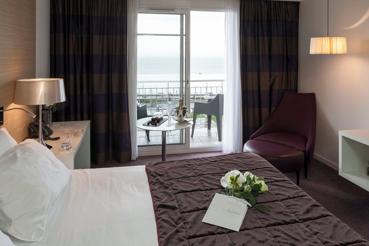 Habitación de hotel con una cama, un escritorio, una silla, una mesa con una botella y vasos, y vista al mar a través de la ventana.