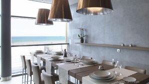 A modern dining area with a view of the ocean, featuring a long table set for dining, pendant lights, and a minimalist decor.
