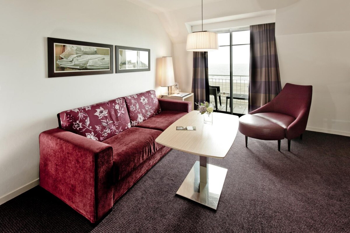 Habitación de hotel con sofá rojo, mesa de centro de madera, sillón de cuero, lámpara y vista al mar a través de una ventana.