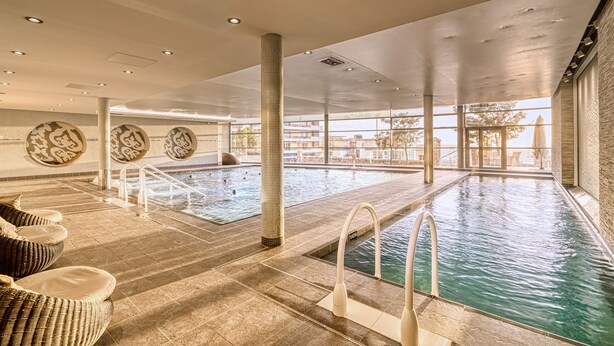 A large indoor swimming pool with clear water, surrounded by lounge chairs and a tiled floor.