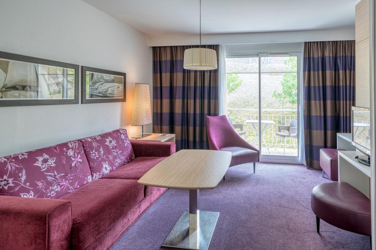 Habitación de hotel con sofá, una mesita, una silla, lámpara y vistas al exterior.
