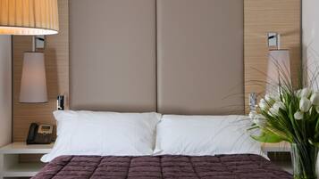 Uma cama bem arrumada com roupa de cama branca e roxa, uma luminária de cabeceira, um telefone e um vaso com flores sobre uma mesa de cabeceira de madeira.