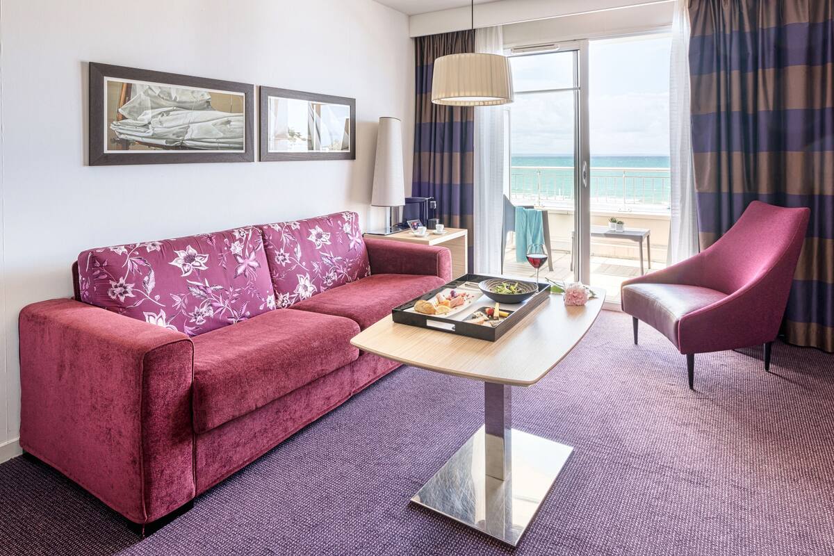 Une chambre d’hôtel avec un canapé rouge, un fauteuil rose, une table à manger sur laquelle se trouve un plateau de nourriture, une lampe et une vue sur la mer par de grandes fenêtres.