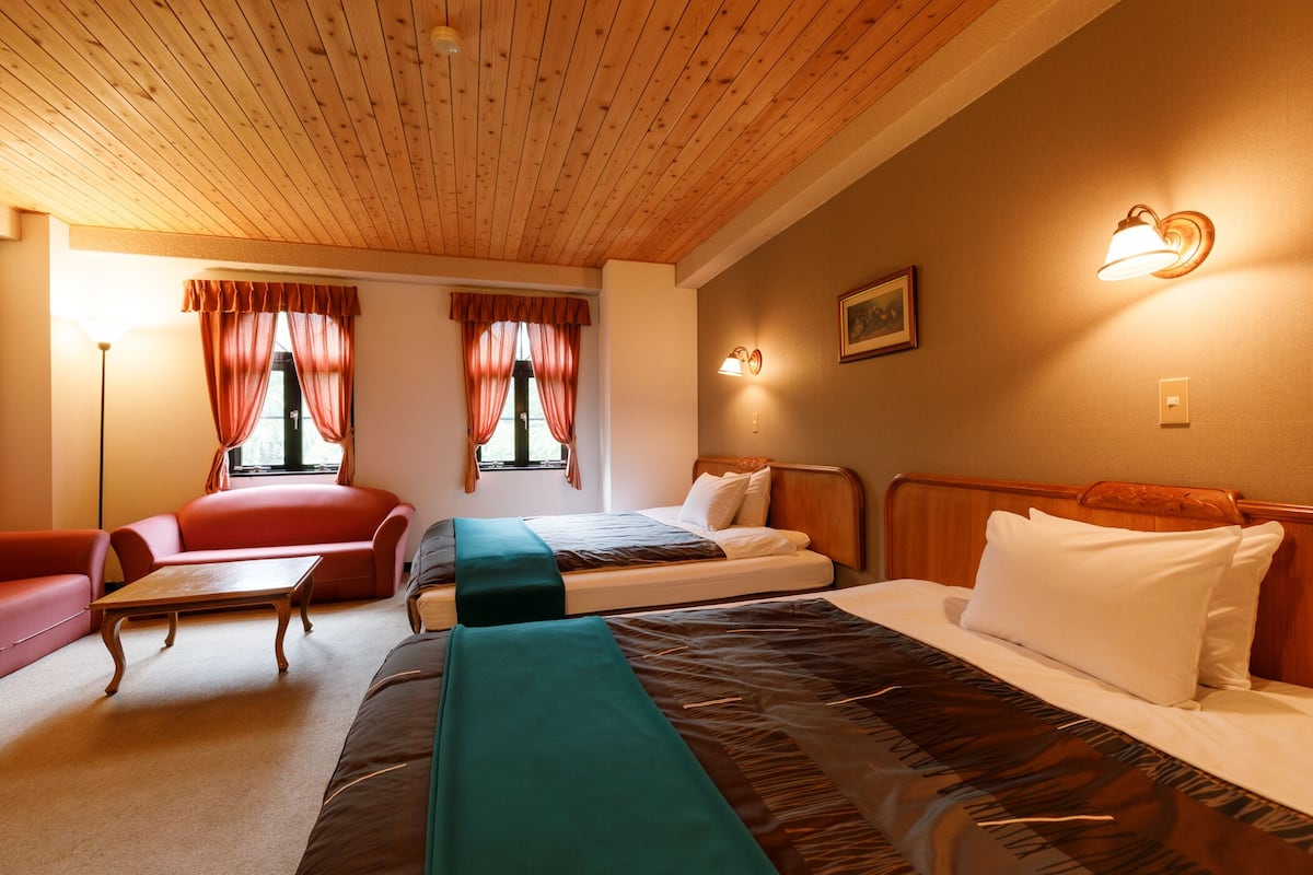 A hotel room with a wooden ceiling, two beds, a sofa, a table, and curtains.
