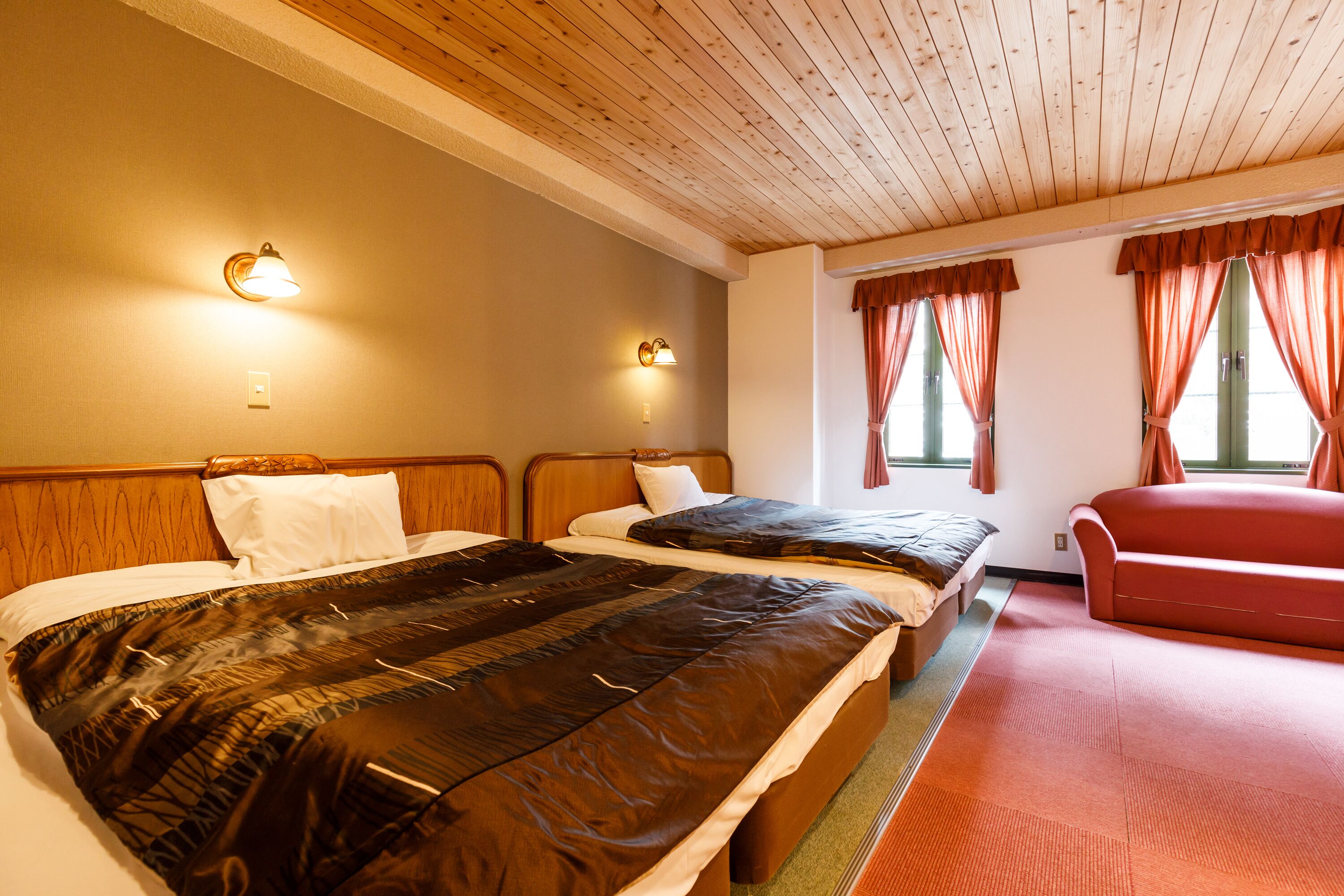 A hotel room with two beds, a red sofa, wooden ceiling, and wall-mounted lights.