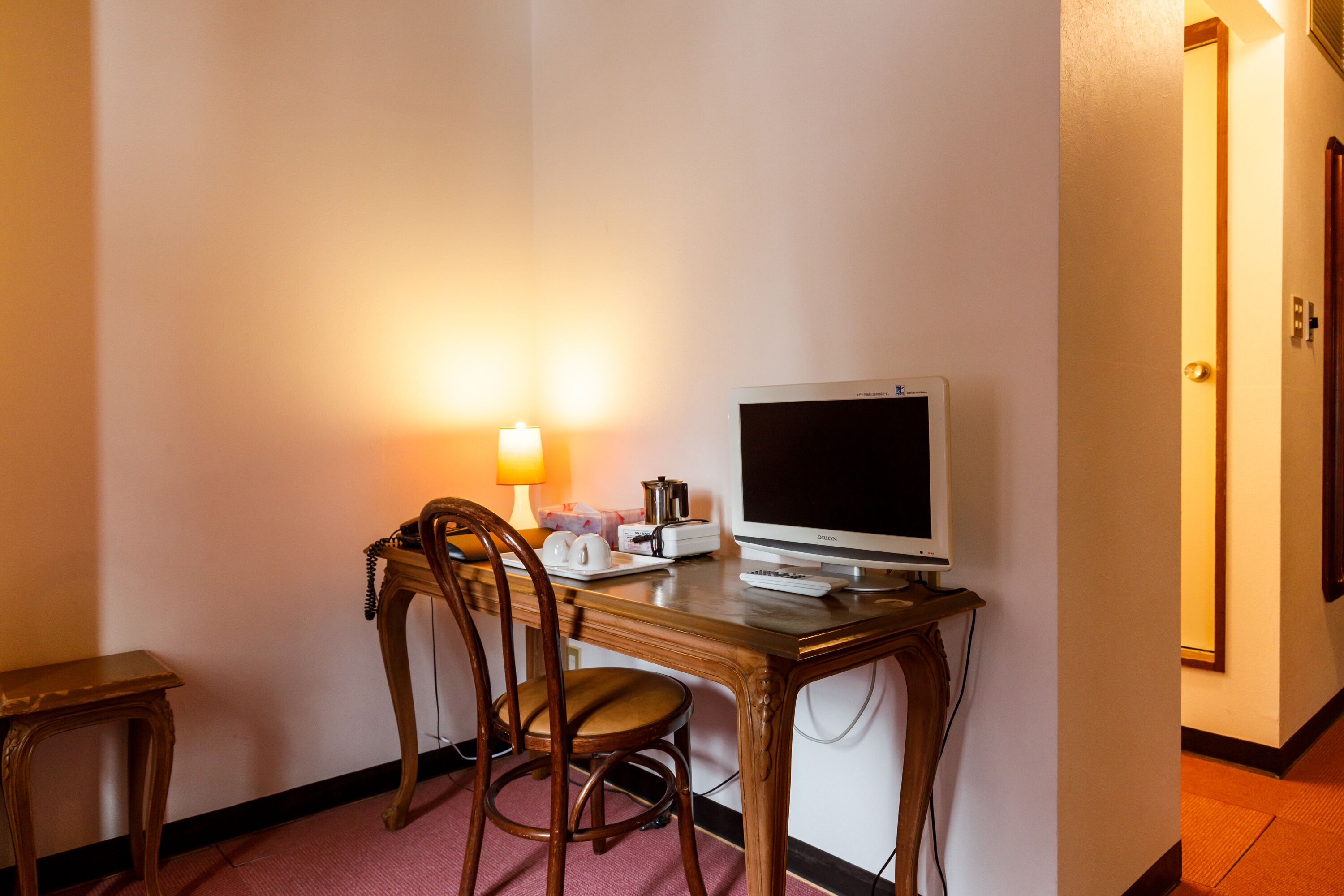A hotel room with a wooden desk, a computer, a chair, a lamp, and a small table with a telephone and a cup.