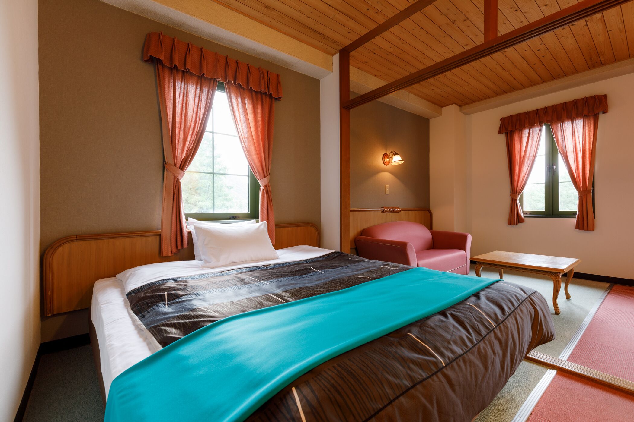 Une chambre d’hôtel avec un grand lit, une tête de lit en bois, une chaise rouge, une petite table et une lampe fixée au mur.