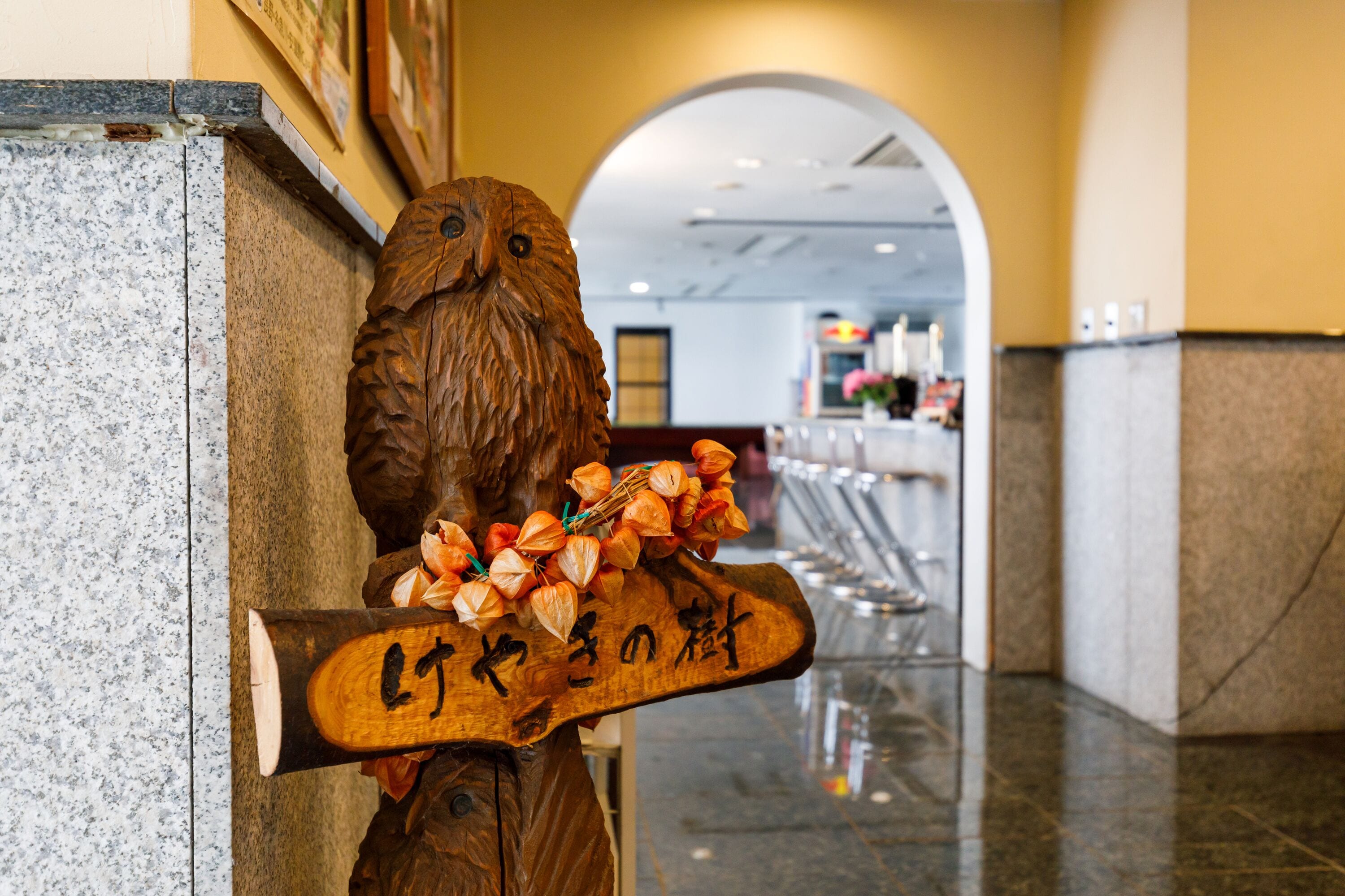 A wooden sculpture of an owl holding a sign with Japanese characters, located in a hallway with a marble floor and an archway in the background.