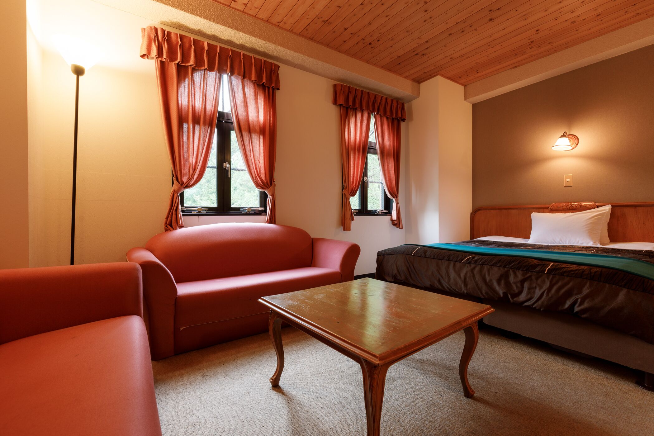 Una habitación con techo de madera, un sofá rojo, una mesa de madera, una cama con manta verde y dos ventanas con cortinas rojas.