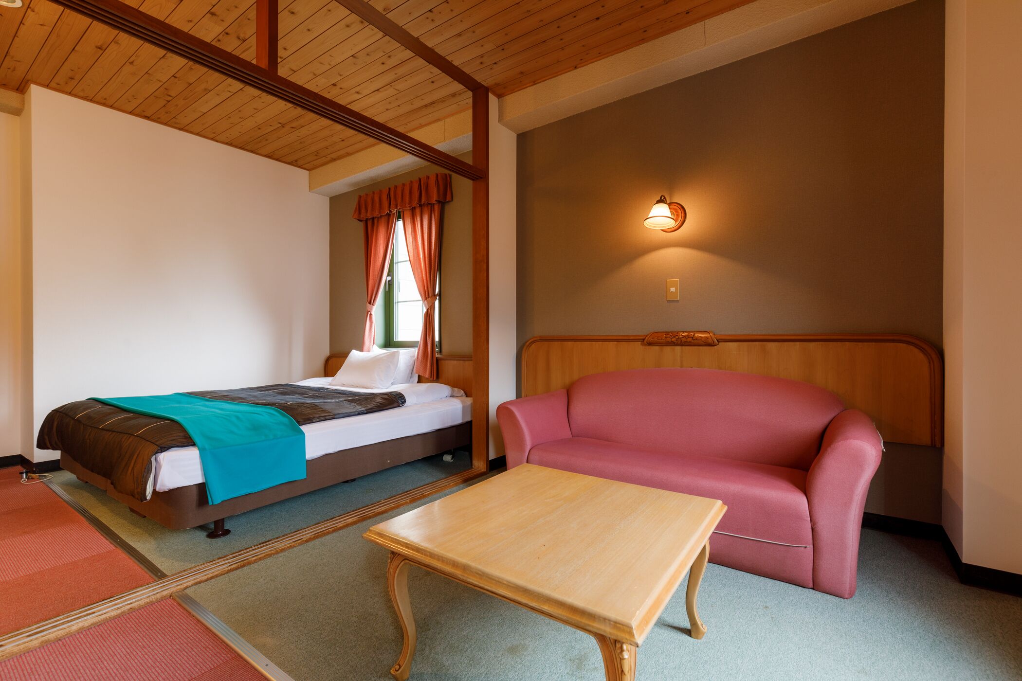 A hotel room with a bed, a red sofa, a wooden table, and a wall-mounted light fixture.