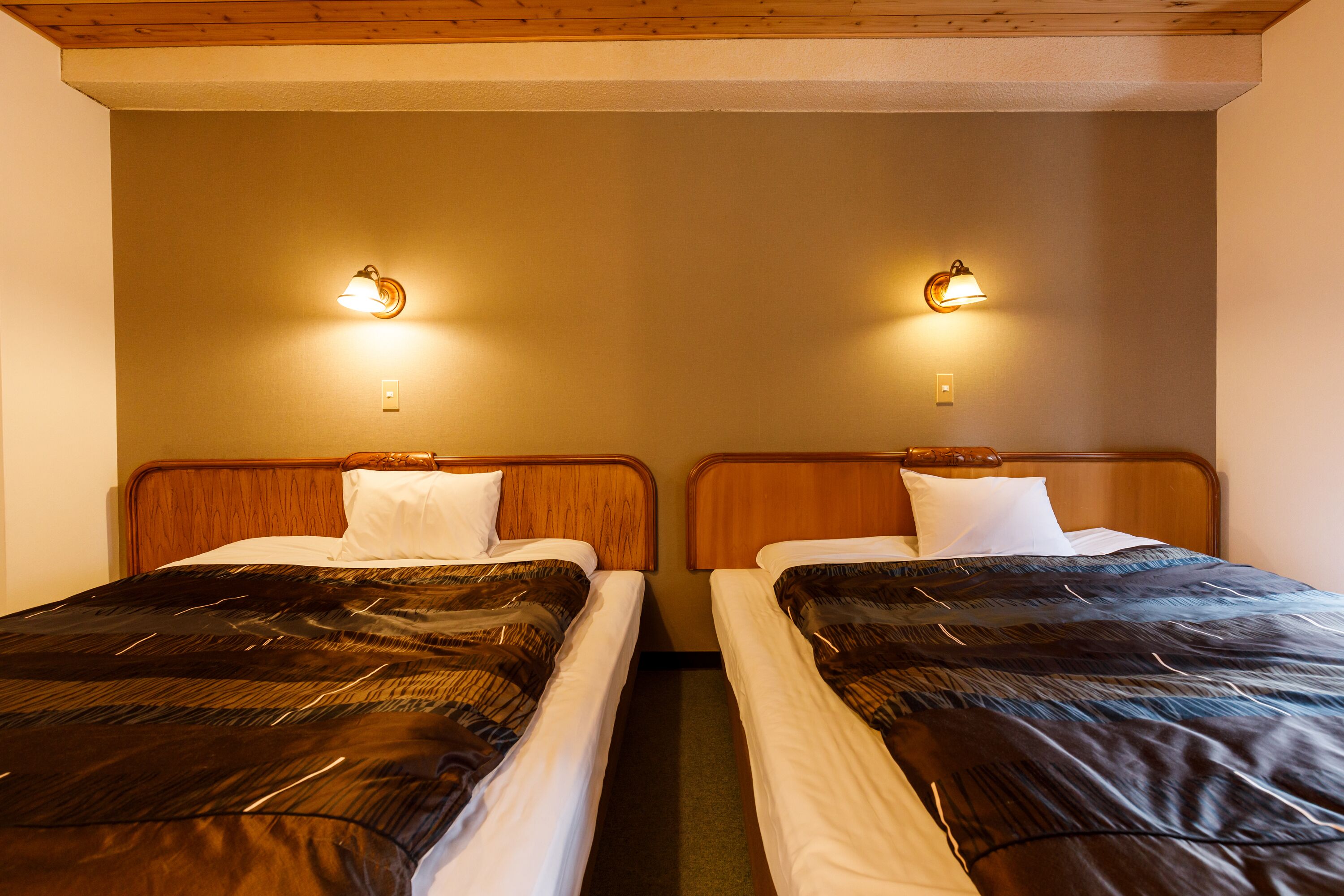 Deux lits simples avec des têtes de lit en bois et une literie blanche, de part et d’autre des appliques murales.