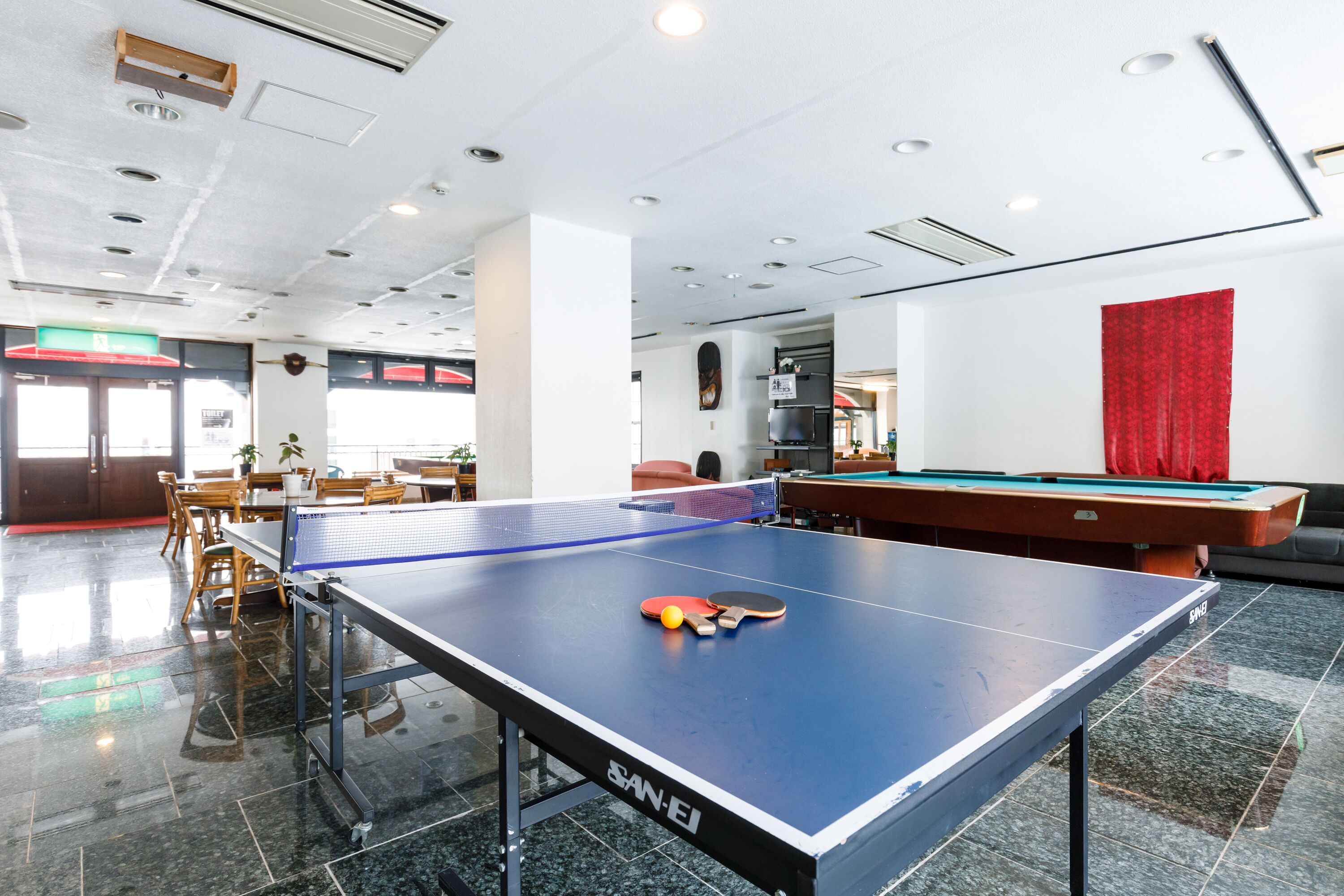 A ping pong table with paddles and a ball in a room with a pool table, chairs, and a red curtain.