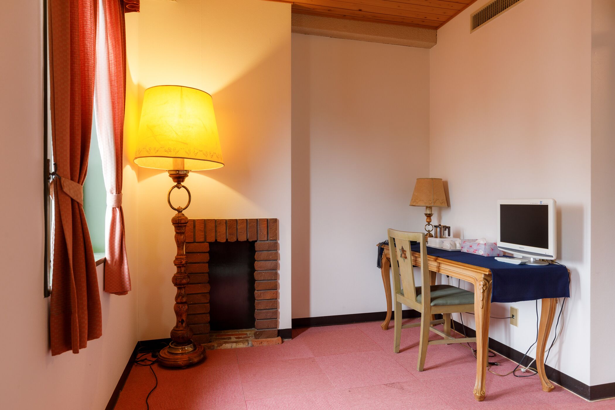 A room with a brick fireplace, a desk with a computer, and a lamp.