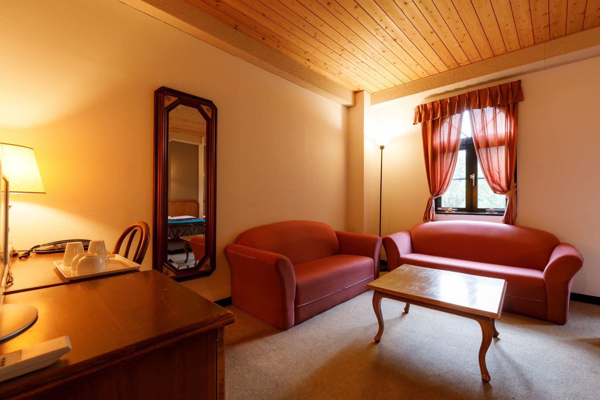 A room with a wooden ceiling, a sofa, a table, a mirror, and a window with curtains.