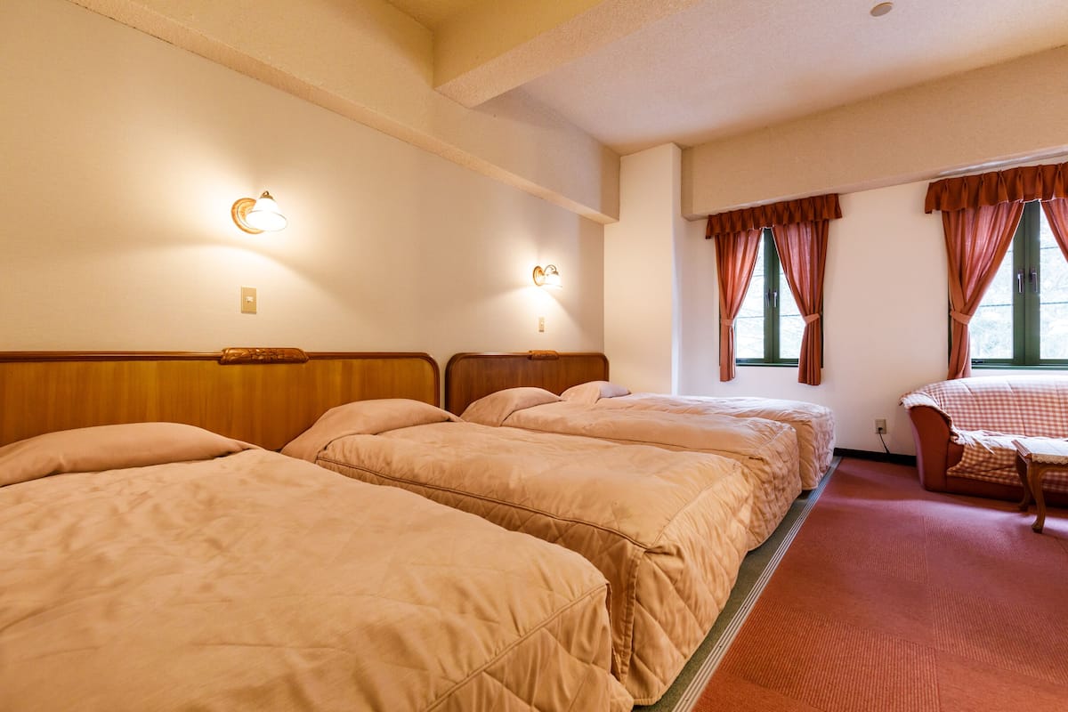 A hotel room with three beds, a wooden headboard, a small table, and two windows with curtains.